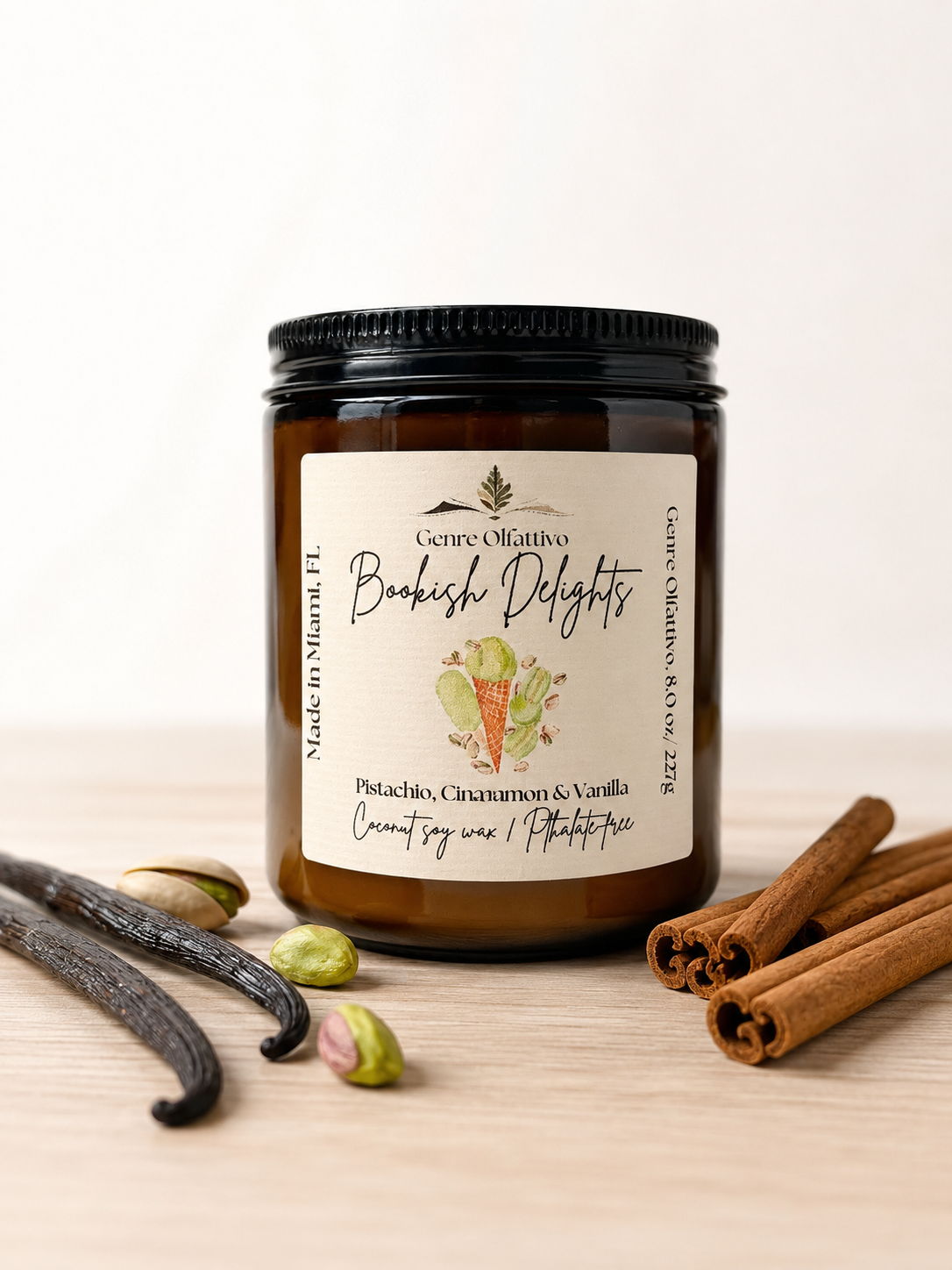 Bookish candle. An amber glass jar candle with a black lid and a cream label reading 'Bookish Delights' and 'Pistachio, Cinnamon & Vanilla' sits on a light wood surface with vanilla beans, pistachios, and cinnamon sticks around it.