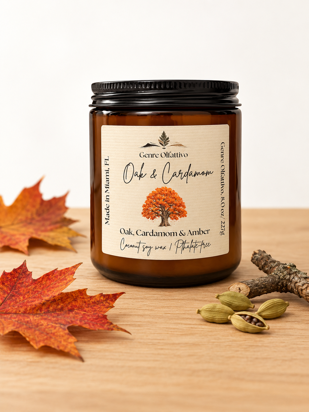 An amber glass jar candle with a black lid and a cream label reading 'Genre Olfattivo Oak & Cardamom,' styled on a wooden surface with autumn leaves, a small branch, and cardamom pods.