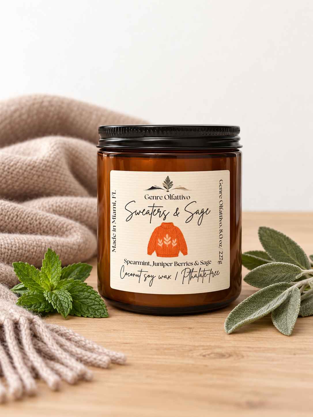 An amber glass jar candle with a black lid and a cream label reading 'Genre Olfattivo' and 'Coconut soy wax / Phthalate free'. The candle is styled on a wooden surface with a beige knit blanket, mint leaves, and sage leaves against a light background.