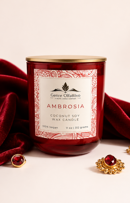 A red glass jar candle with a gold lid and a cream label reading Genre Olfattivo Ambrosia Coconut Soy Wax Candle, BR540 dupe, styled with draped red velvet fabric and gold jewelry on a light 
beige background.