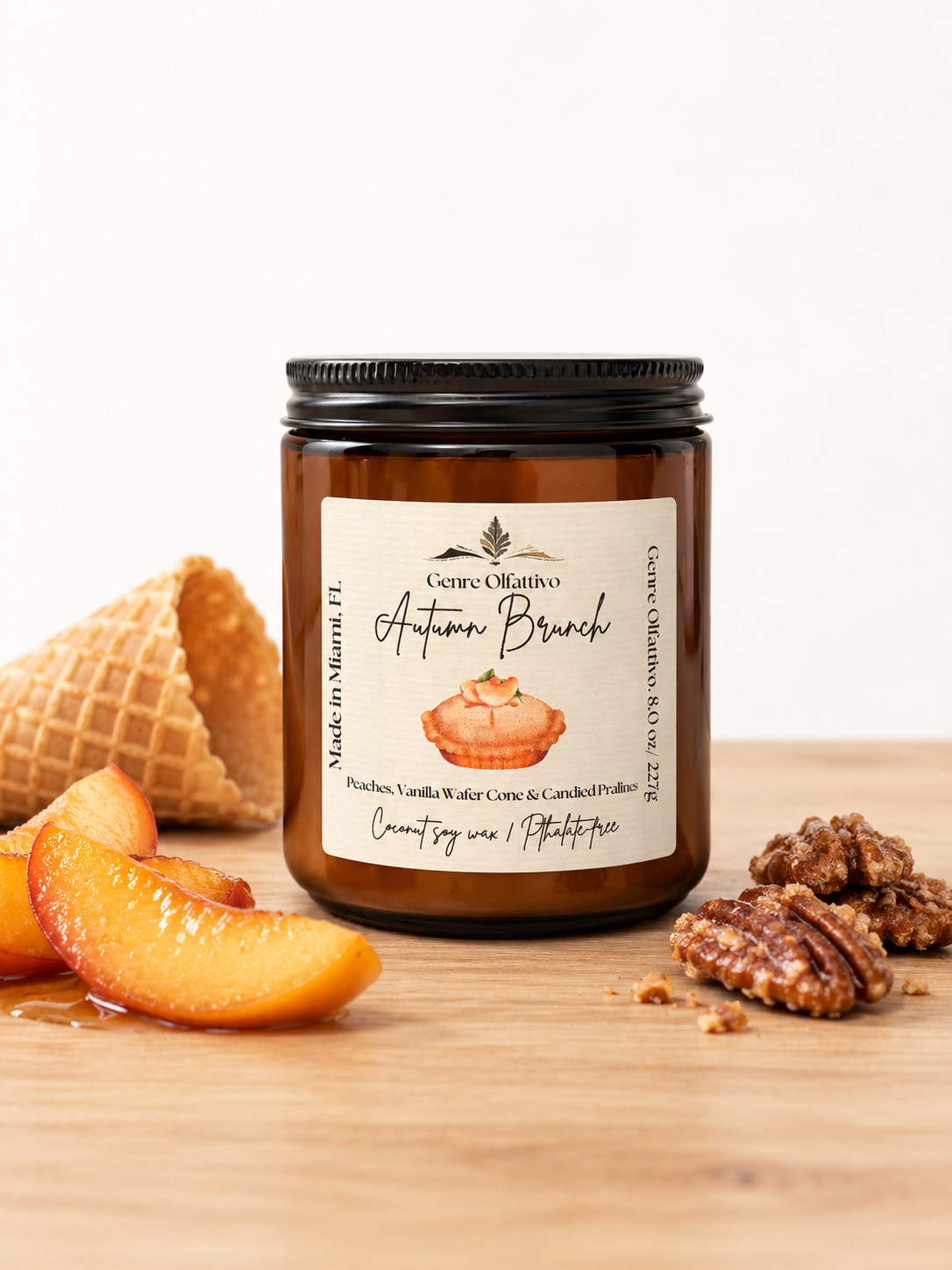 An amber glass jar candle with a black lid and a cream label reading 'Autumn Brunch' by Genre Olfattivo Candles, styled on a wooden surface with peach slices, a waffle cone, and pecans against a white background.