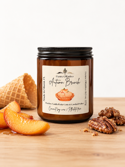 An amber glass jar candle with a black lid and a cream label reading 'Autumn Brunch' by Genre Olfattivo Candles, styled on a wooden surface with peach slices, a waffle cone, and pecans against a white background.