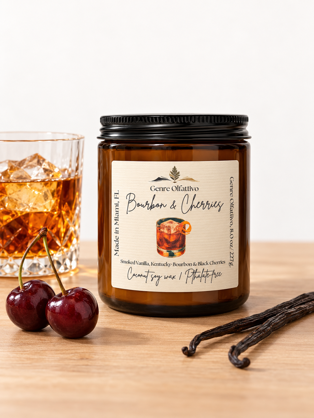 A candle jar labeled 'Genre Olfattivo’ Bourbon & Cherries' sits on a wooden surface beside a glass of amber drink, two cherries, and vanilla beans. The candle is in an amber glass jar with a black lid and cream label.