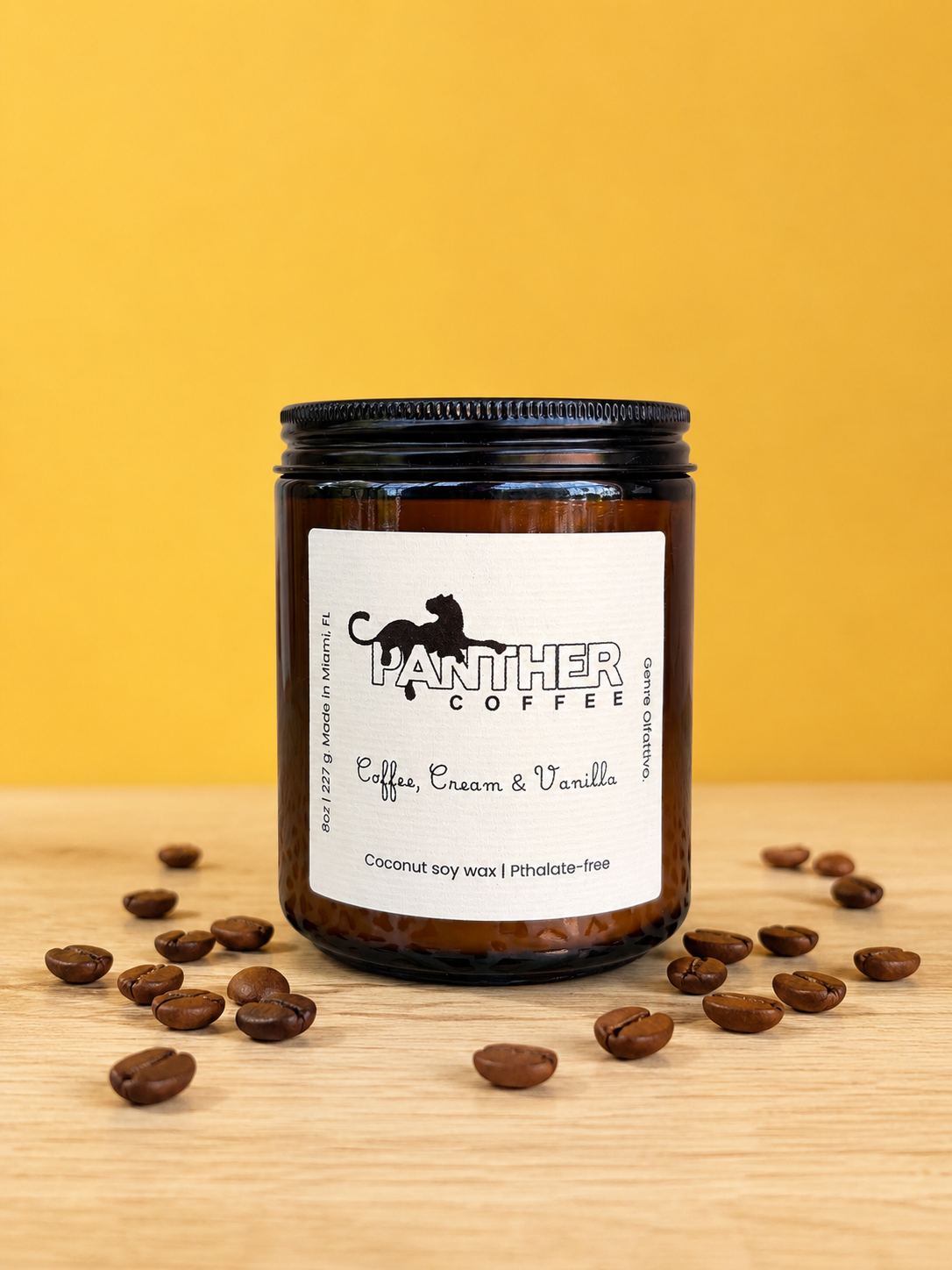 A dark amber glass jar of Panther Coffee 'Coffee, Cream & Vanilla' soy wax candle sits on a light wood surface with scattered coffee beans, set against a solid yellow background.