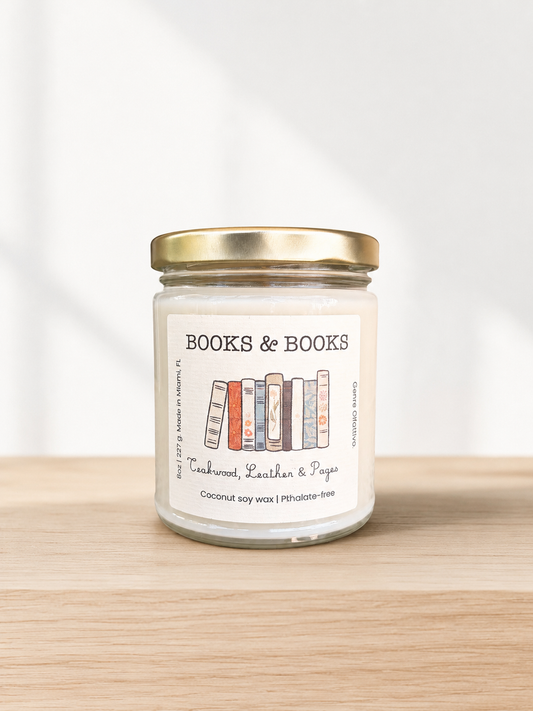 A glass jar candle with a gold metal lid and a cream-colored label reading 'BOOKS & BOOKS' with an illustration of colorful books, set on a light wood surface against a white background.