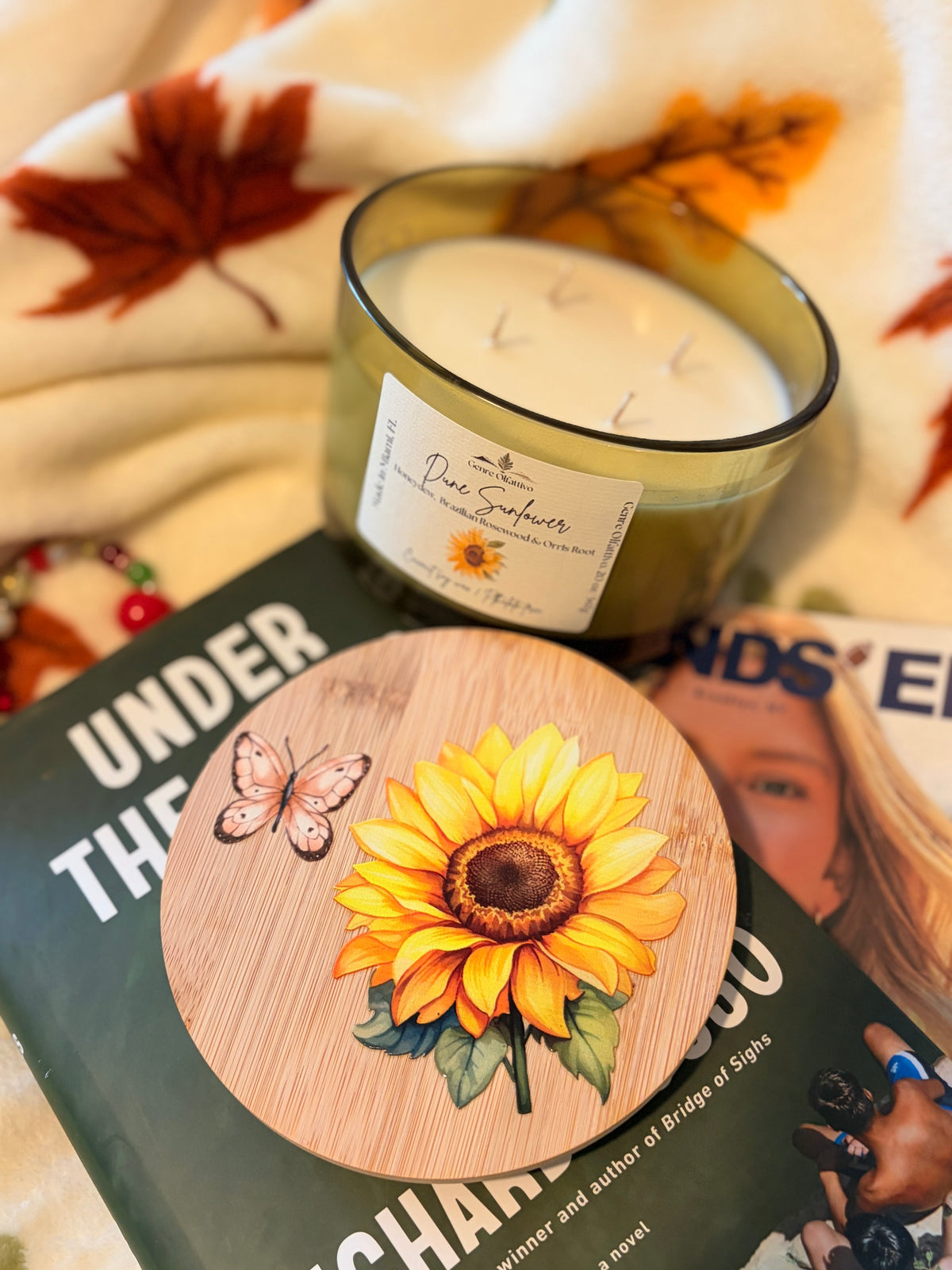 A round wooden coaster featuring a painted yellow sunflower and butterfly, styled on top of a book next to a candle in a glass jar with a cream label.
