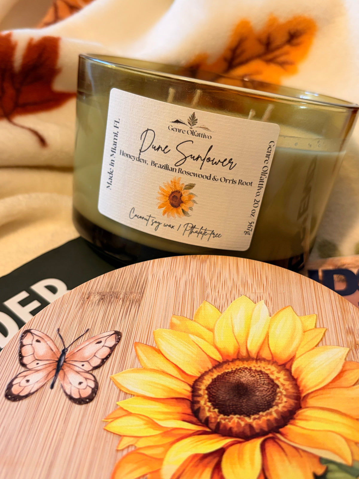 A candle in a dark green-tinted glass jar with a white label reading 'Dune Sunflower' by Genre Olfattivo, styled on a wooden surface with a matching sunflower coaster and autumn leaves in the background.