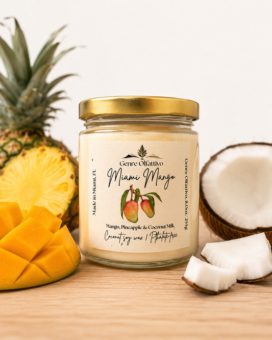 A glass jar candle with a gold lid and cream-colored label reading 'Genre Olfattivo Miami Mango', styled on a wooden surface with mango slices, pineapple, and coconut halves around it.