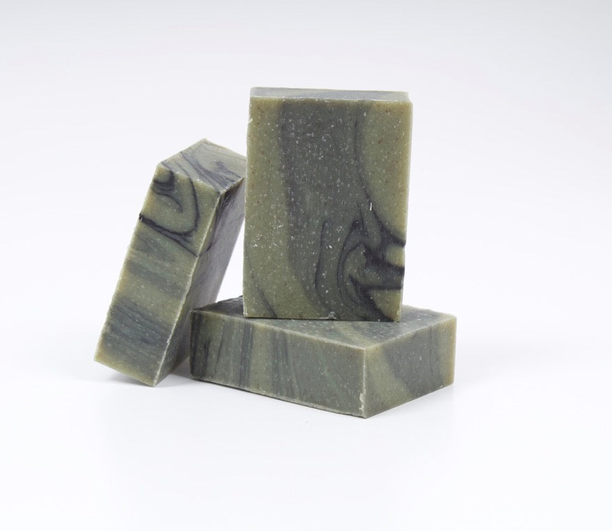 Three rectangular bars of soap stacked and leaning on a white studio background. The soap has a muted olive green base with dark gray to black marbled swirls. Cold pressed soap, Fraser Fir and Pine scented, Christmas Soap. 