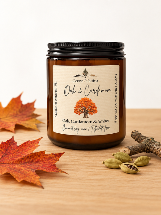 An amber glass jar candle with a black lid and a cream label reading 'Genre Olfattivo Oak & Cardamom,' styled on a wooden surface with autumn leaves, a small branch, and cardamom pods.
