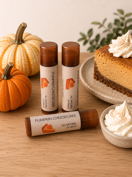 Three lip balm tubes labeled 'Pumpkin Cheesecake' arranged on a wooden table with autumn props, including pumpkins, a slice of pumpkin pie, and whipped cream.