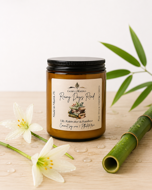 An amber glass jar candle with a black lid and a cream label reading 'Rainy Days Read’ by Genre Olfattivo, styled on a light wood surface with white flowers and green bamboo leaves.