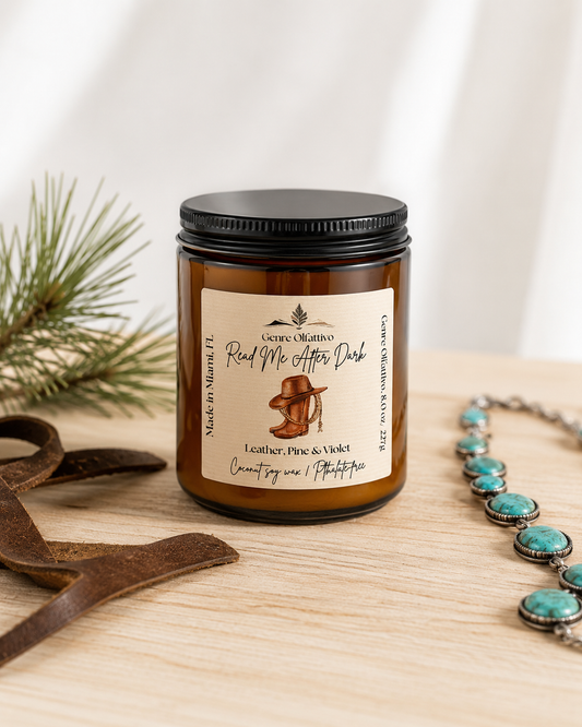 An amber glass jar candle with a black lid and a cream label reading 'Read Me After Dark’, styled on a light wood surface with a turquoise necklace, leather ribbon, and pine branches.